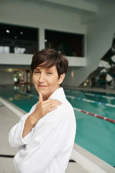 Feliz mulher de meia-idade com cabelo curto em roupão branco, centro de spa, piscina interior, sorriso — Fotografia de Stock