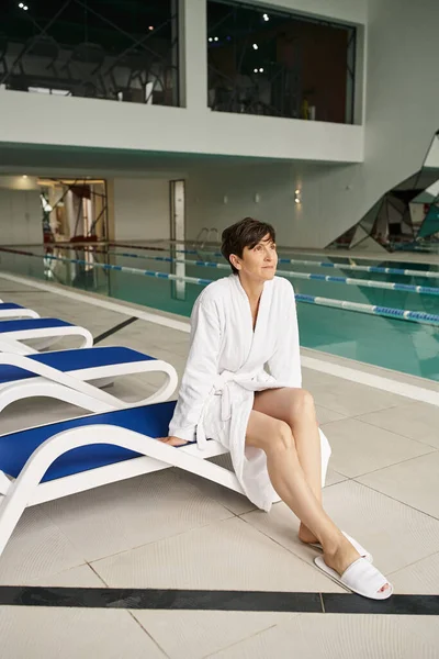 Frau mittleren Alters mit kurzen Haaren sitzt auf einer Liege, weißer Bademantel, Wellness-Center, innen, Schwimmbad — Stockfoto