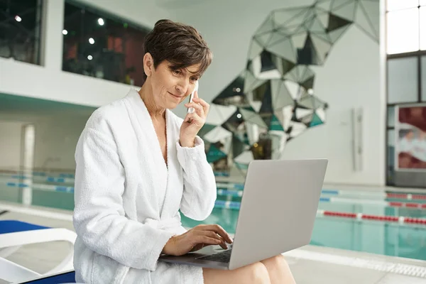 Freelancer, middle aged woman talking on smartphone, using laptop, sitting on lounger, spa center — Stock Photo