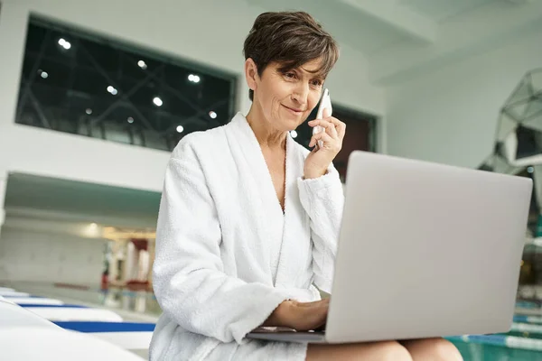 Freelance, phone call, middle aged woman talking on smartphone, laptop, lounger, spa center — Stock Photo