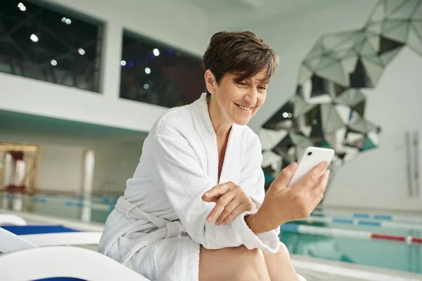 Centro de spa, mujer de mediana edad feliz con teléfono inteligente, sentado en la tumbona, bata blanca, piscina - foto de stock