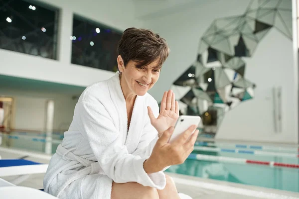 Centre de spa, heureuse femme d'âge moyen ayant un appel vidéo sur smartphone, chaise longue, peignoir, piscine — Photo de stock