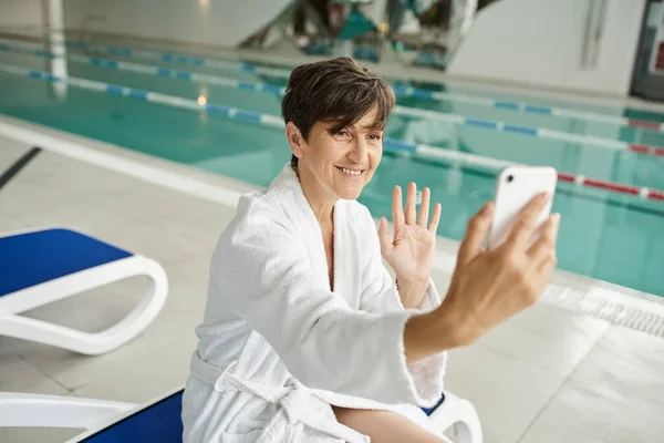 Mujer de mediana edad positiva que tiene videollamada en el teléfono inteligente, tumbona, piscina, centro de spa - foto de stock