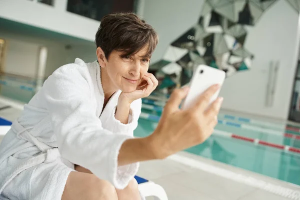 Alegre mujer de mediana edad en bata blanca tomando selfie, teléfono inteligente, centro de spa, piscina - foto de stock