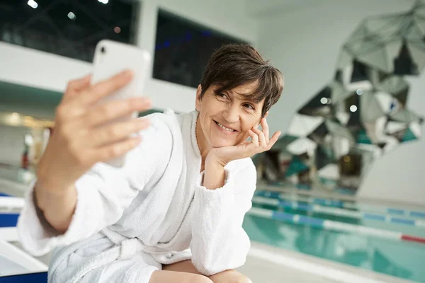 Cheerful middle aged woman in white robe taking selfie near swimming pool, smartphone, spa center — Stock Photo