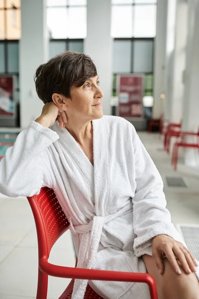 Pose relajada, mujer madura soñadora en bata blanca sentada en silla roja, interior, centro de spa, mirar hacia otro lado - foto de stock