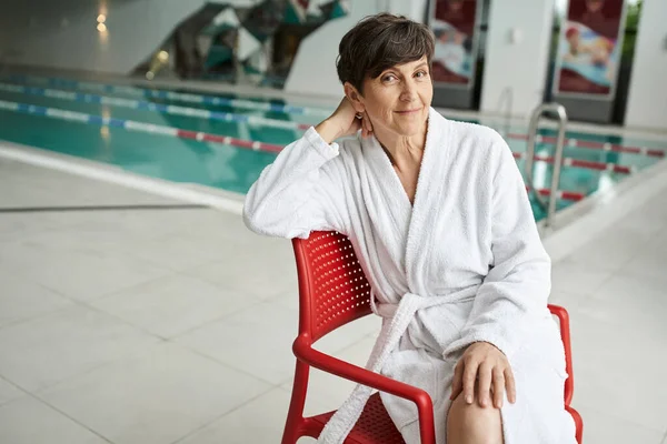 Posa rilassata, felice donna matura in accappatoio bianco seduta sulla sedia rossa, piscina coperta, giornata termale — Foto stock