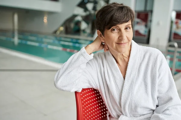 Pose relaxada, mulher madura alegre no manto branco que senta-se na cadeira vermelha, piscina interna, dia do spa — Fotografia de Stock