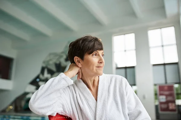 Relaxed pose, joyful mature woman in white robe sitting on red chair, indoor spa center, tranquil — Stock Photo