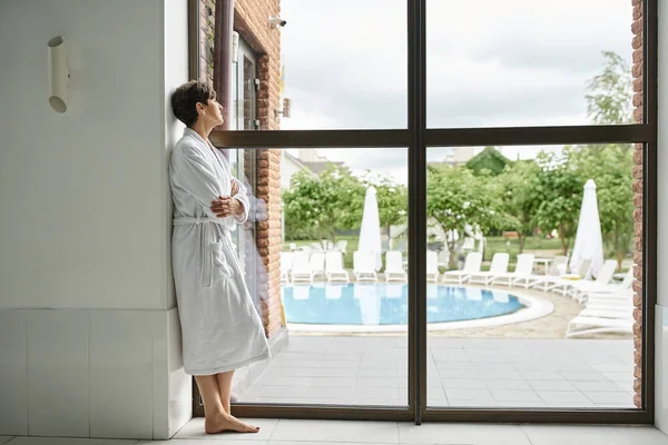 Middle aged woman with short hair standing in white robe near panoramic window in spa center, pool — Stock Photo