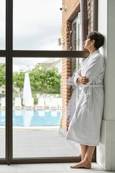 Donna matura con capelli corti in piedi in accappatoio bianco vicino alla finestra panoramica nel centro benessere, vista piscina — Foto stock