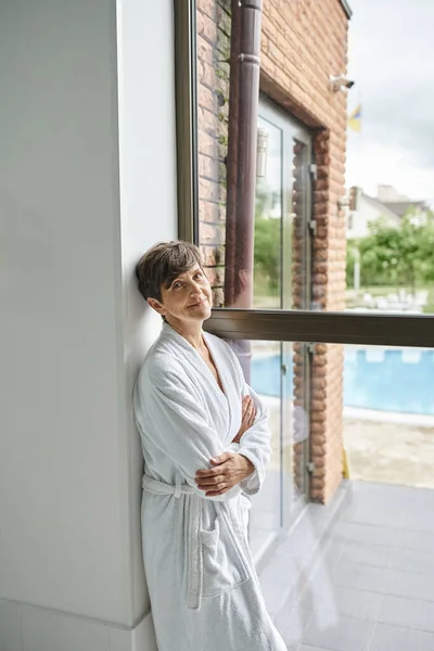 Femme mature debout en robe près de la fenêtre panoramique dans le centre spa, vue sur la piscine, regardez la caméra — Photo de stock