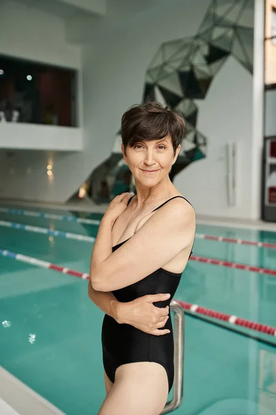 Heureuse femme d'âge moyen avec les cheveux courts debout en maillot de bain près de la piscine intérieure, centre de spa, sport — Photo de stock