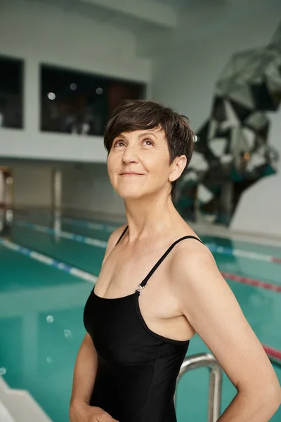 Мечтательная женщина среднего возраста с короткими волосами позирует в купальнике рядом с крытым бассейном, спа-центр, смотреть вверх — стоковое фото