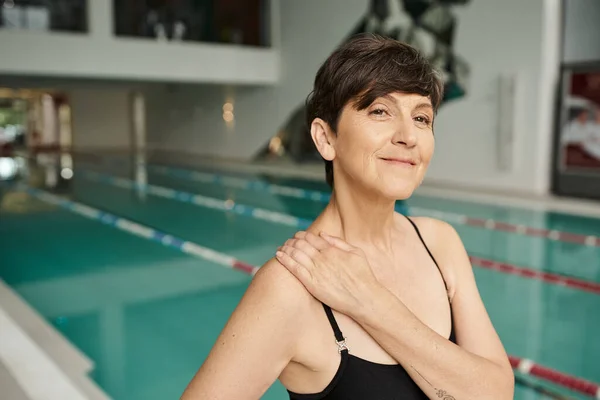 Feliz e madura, mulher tatuada, esportista no centro de spa, olhando para longe, piscina, roupa de banho — Fotografia de Stock