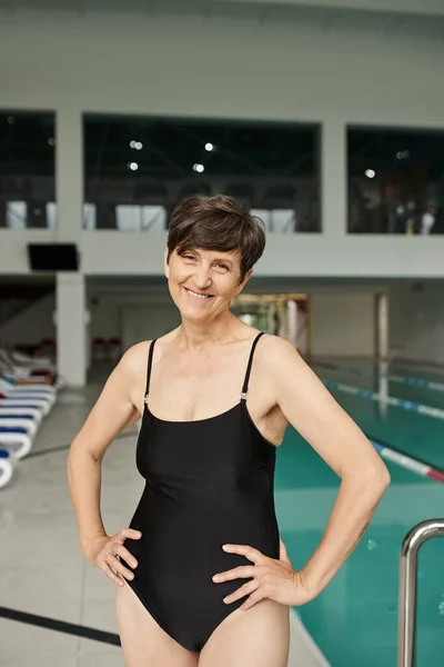 Positiva donna di mezza età in piedi con le mani sui fianchi vicino alla piscina, costumi da bagno, centro benessere — Foto stock
