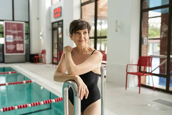 Femme d'âge moyen rêveuse en maillot de bain posant près de la piscine intérieure, centre de spa, portrait — Photo de stock