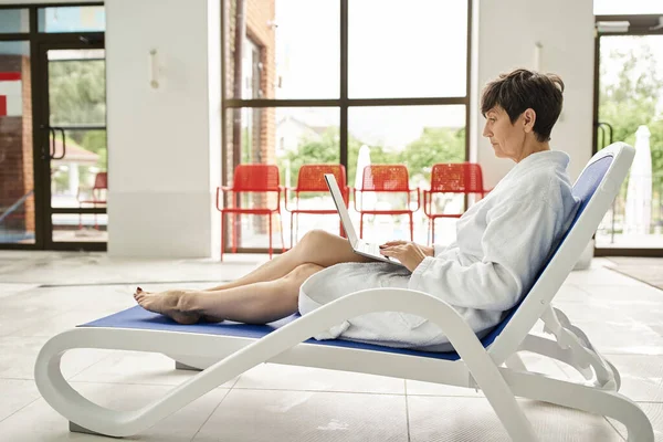 Freelance e spa, mulher de meia-idade em roupão branco usando laptop, sentado na espreguiçadeira, trabalho remoto — Fotografia de Stock