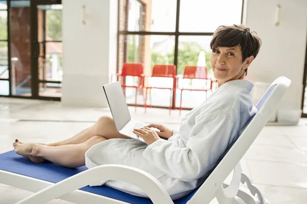 Heureuse femme d'âge moyen en robe blanche à l'aide d'un ordinateur portable, assise sur une chaise longue, télétravail, centre de spa — Photo de stock