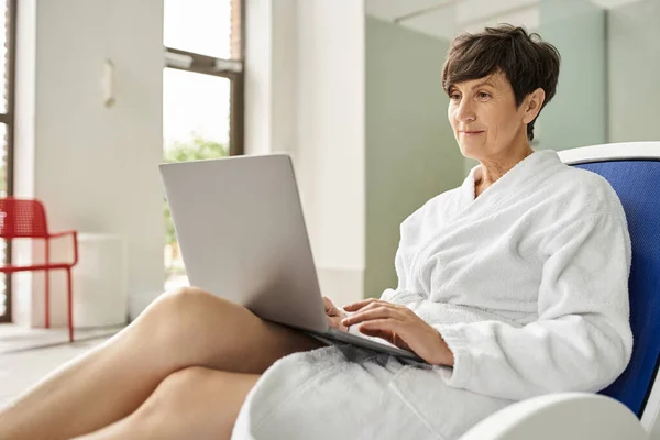 Middle aged woman in white robe using laptop, sitting on lounger in spa center, remote work, indoor — Stock Photo