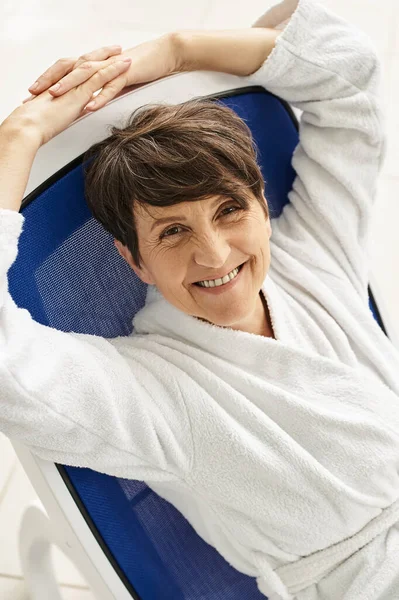 Happy mature woman with short hair relaxing on lounger, white robe, spa center, look at camera — Stock Photo