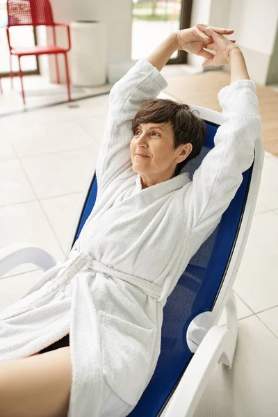 Joyful middle aged woman with short hair stretching on lounger, white robe, spa center, smile — Stock Photo