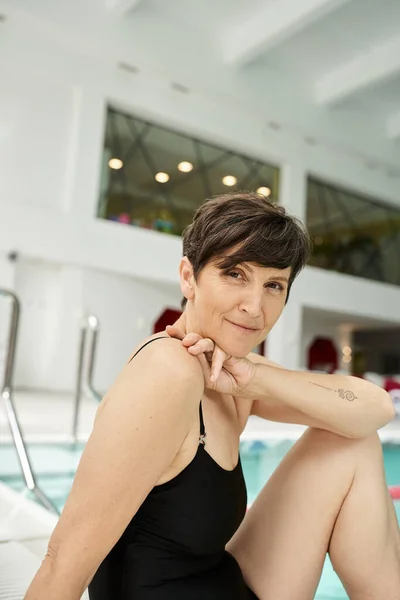 Retrato de mujer de mediana edad tatuada en traje de baño sentado cerca de la piscina, junto a la piscina, centro de spa - foto de stock