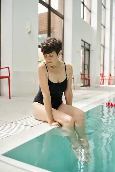 Mujer madura en traje de baño sentado junto a la piscina con las piernas en el agua, piscina, centro de spa - foto de stock
