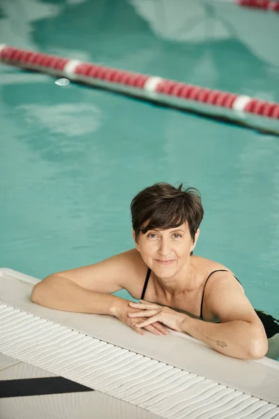 Mujer de mediana edad feliz con tatuaje nadando en la piscina, en el interior, centro de spa, bienestar, relajación - foto de stock