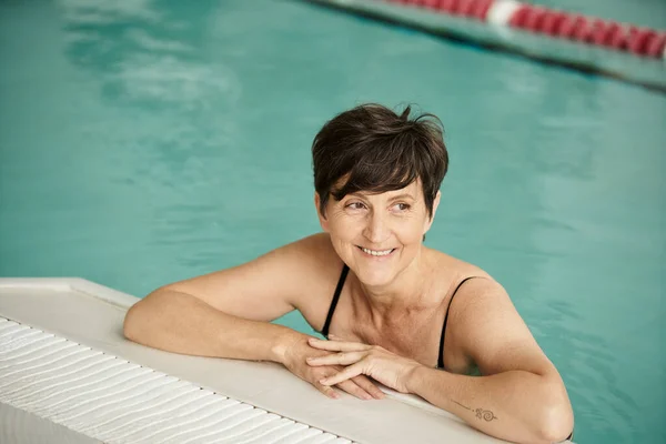 Mujer alegre de mediana edad con tatuaje nadando en la piscina, en el interior, centro de spa, bienestar, relajación - foto de stock