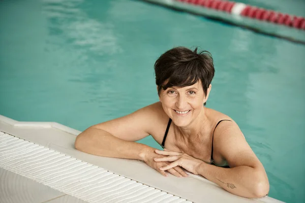 Mujer alegre y madura con tatuaje nadando en la piscina, en el interior, centro de spa, bienestar, relajación - foto de stock