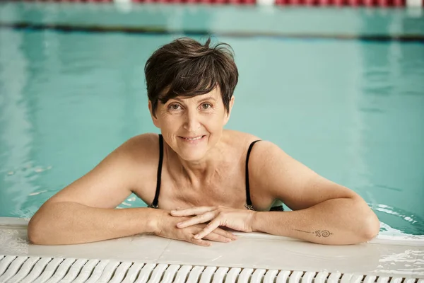 Tatuata donna di mezza età che nuota in piscina, capelli corti, guarda telecamera, indoor, centro benessere — Foto stock