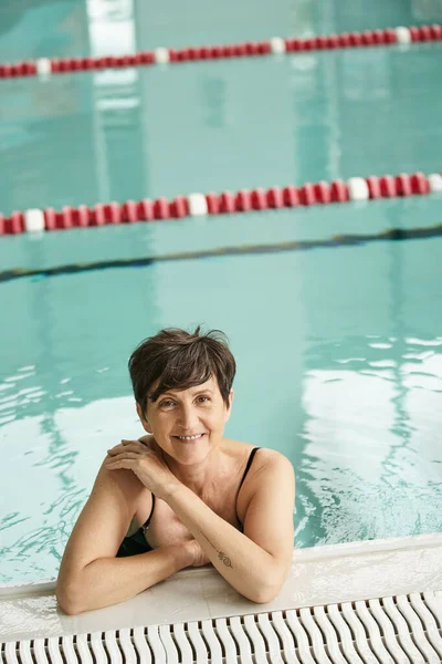 Mujer positiva de mediana edad con pelo corto, junto a la piscina, mira a la cámara, centro de spa interior, nadar - foto de stock