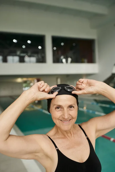 Joyful middle aged woman in goggles and swim cap, swimmer in wellness center, swimming pool — Stock Photo