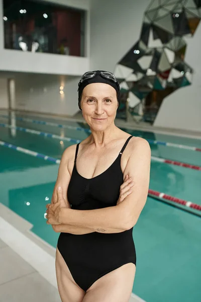 Tattooed middle aged woman in swim cap and goggles, swimming pool in wellness center, folded arms — Stock Photo