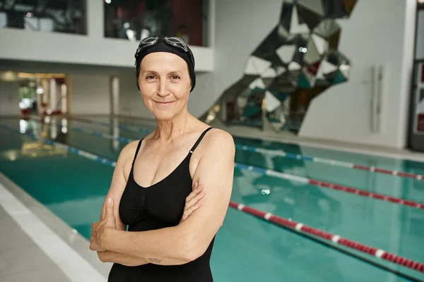 Cheerful middle aged woman in swim cap and goggles, swimming pool in wellness center, folded arms — Stock Photo