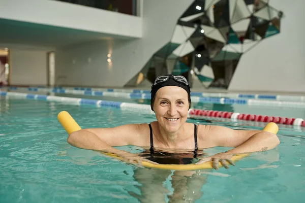 Cheerful middle aged woman in swim cap and goggles swimming with pool noodle, healthy lifestyle — Stock Photo