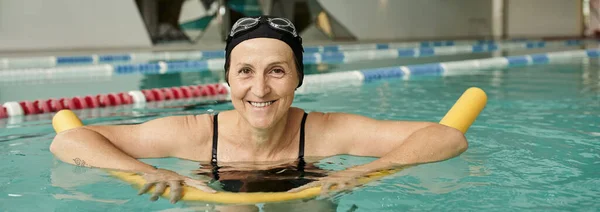 Щаслива зріла жінка в шапці для плавання та окулярах плаває з локшиною для басейну, здоровий спосіб життя, банер — стокове фото