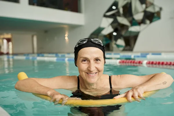 Happy mature woman in swim cap and goggles swimming with pool noodle, healthy lifestyle, sport — Stock Photo