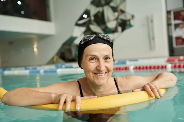 Happy middle aged woman in swim cap and goggles swimming with pool noodle, healthy lifestyle, sport — Stock Photo