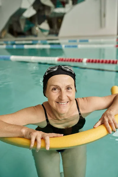 Happy middle aged woman swimming with pool noodle, swim cap and goggles, healthy lifestyle, sport — Stock Photo