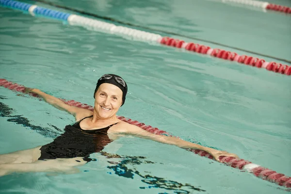 Allegra donna di mezza età che nuota in piscina, sorriso, cuffia e occhiali da bagno, stile di vita sano, sport — Foto stock