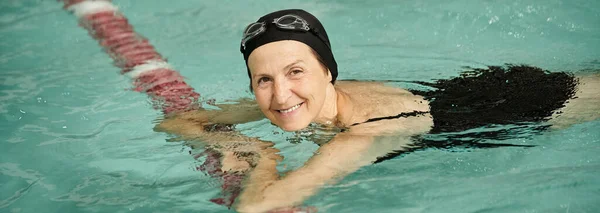 Mujer positiva de mediana edad nadando en la piscina, gorra de natación y gafas, mirando a la cámara, deporte, pancarta - foto de stock