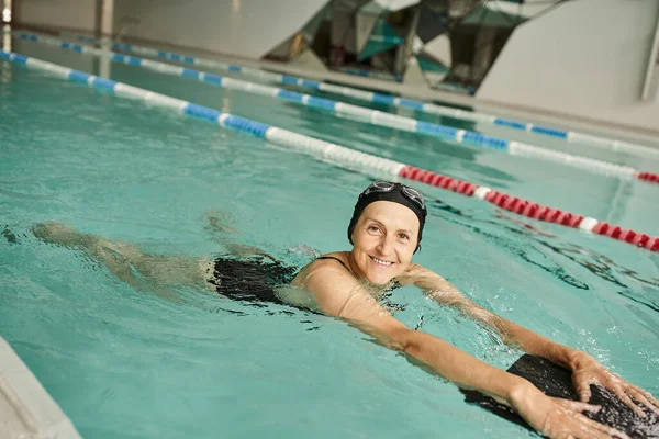 Felice donna di mezza età nuoto con bordo galleggiante in piscina, cuffia e occhiali da bagno, stile di vita — Foto stock