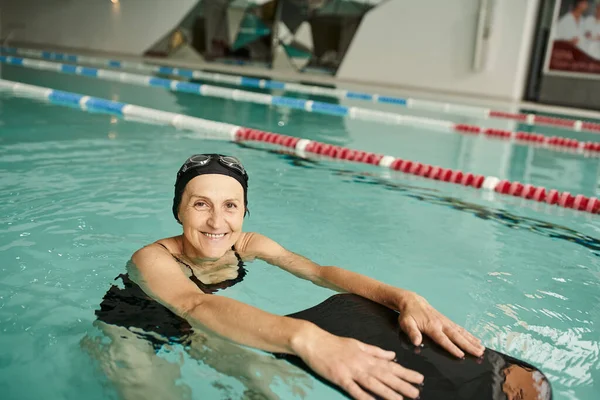 Mulher feliz nadando com prancha flutuante na piscina, touca de natação e óculos, sorriso, meia-idade — Fotografia de Stock