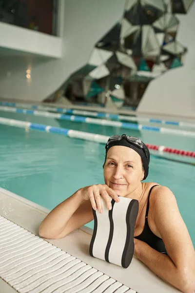 Щаслива жінка середнього віку в шапці для плавання та окулярах біля басейну, посмішка, піна подушка, центр відпочинку — стокове фото