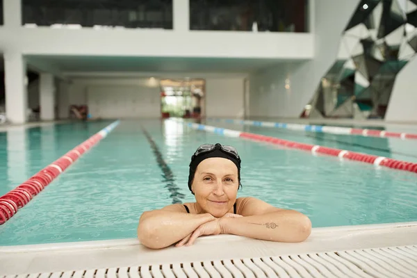 Alegre mulher de meia-idade relaxante à beira da piscina, touca de natação e óculos, sorriso, centro de recreação — Fotografia de Stock