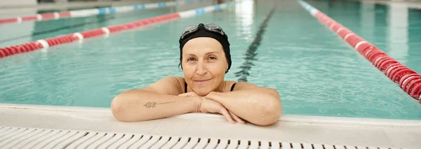 Cheerful middle aged woman relaxing at poolside, swim cap and goggles, recreation center, banner — Stock Photo