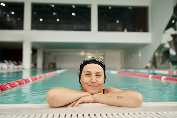 Sportliche Frau mittleren Alters entspannt sich am Pool, Badekappe und -brille, Lächeln, Erholungszentrum — Stockfoto