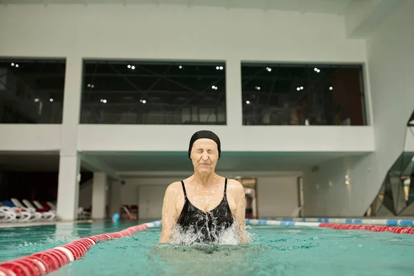 Salpicaduras de agua, mujer madura en gorra de baño y gafas que se divierten en la piscina, ojos cerrados, centro de spa - foto de stock